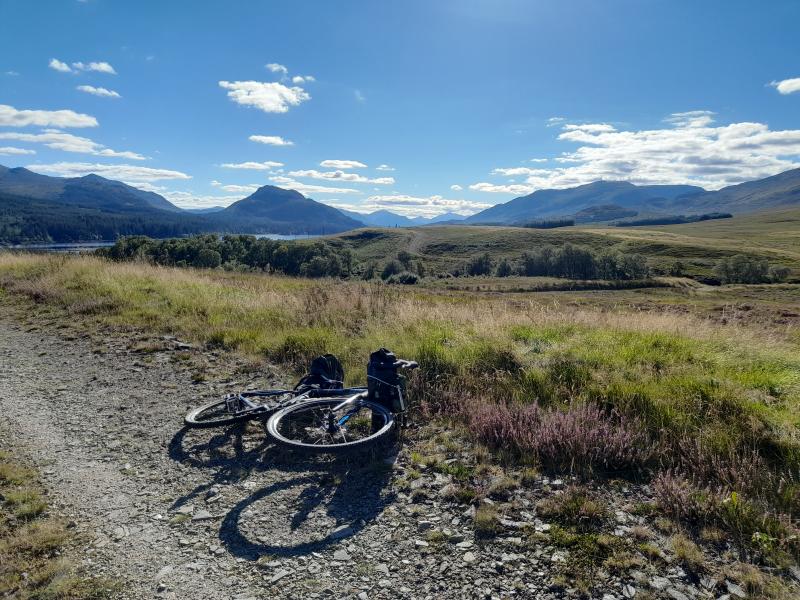 A bicycle lays on the ground in front of a lush green landscape, a lake and mountain range in the background