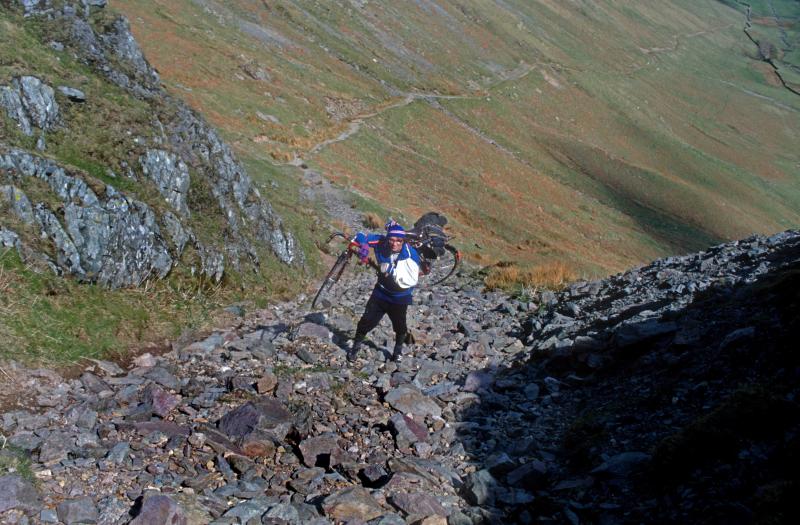 A cyclist with a broken arm carries his bike up a stoney track