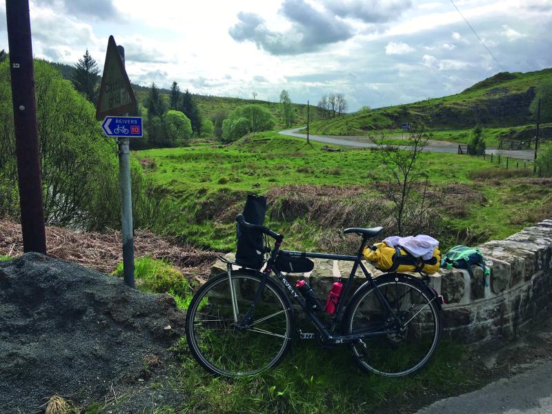 Alf's bicycle leans against a stone wall above a river. In the background there is a snaking road between hilly grassland and trees. In the foreground a roadsign pointing left reads 'Reivers' next to a bicycle symbol