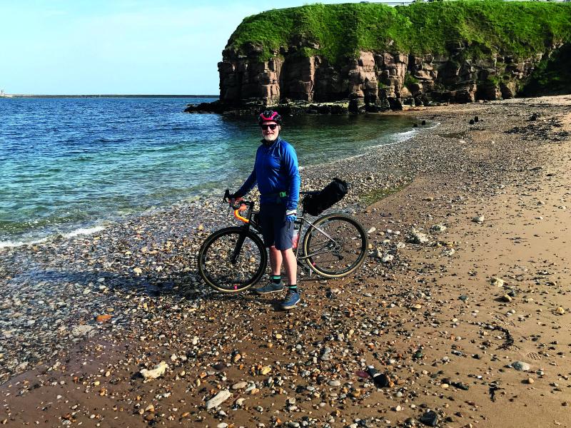 Alf stands with his bicycle on a beach in a cove with a cliff behind him, the blue sea to the left of him