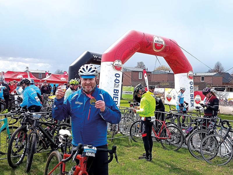 A man wearing a cycle helmet and jacket holds up a medal. He's stood with his bicycle at a finish line