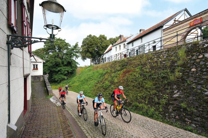 Cyclists ride along a cobbled street flanked by houses either side