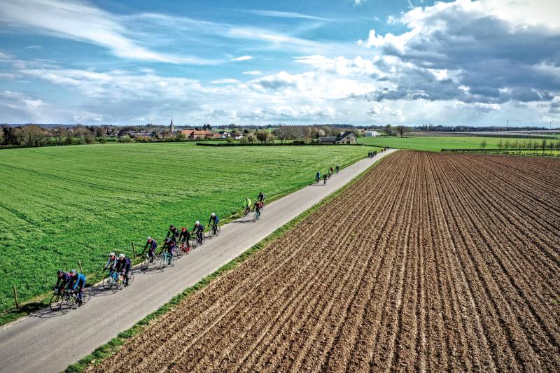 Several cyclists ride along a long straight road between two flat fields, houses and a church in the background