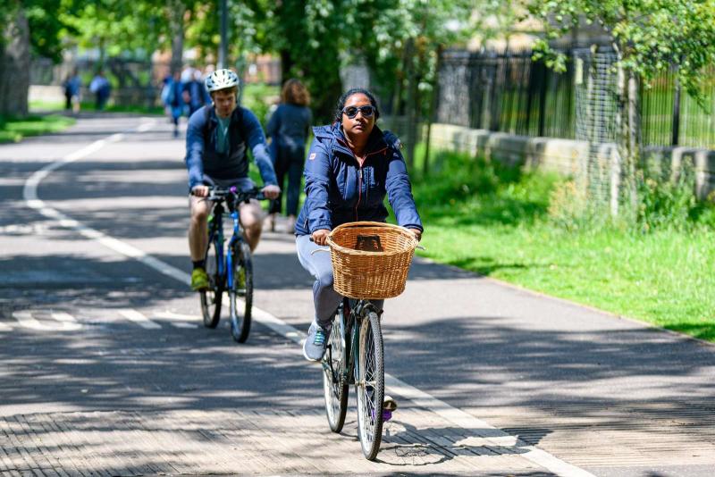 Two people are cycling along a cycle path. They are both wearing normal clothes. The path runs alongside a pavement and there are people walking in the background.