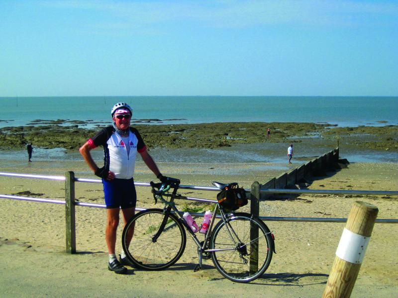 An older man stands by the beach holding his road bicycle next to him. He is wearing cycling shorts and a short-sleeve jersey
