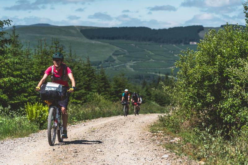 Three people climb a gravel track through a forest