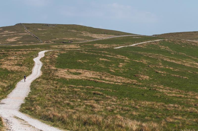 A gravel track curves around a hillside, with a lone person cycling along it