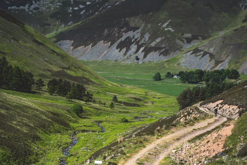 A steep sided mountain valley with a small stream meandering through the middle. A cyclist rides along a gravel track on one side