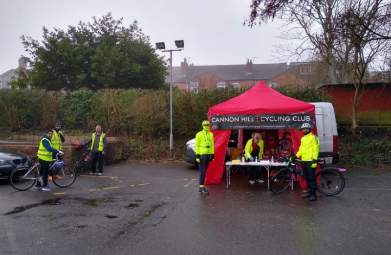 A distant shot of a people under a gazebo on a wet looking day, two cyclists in yellow jackets appear to be heading to visit the stand