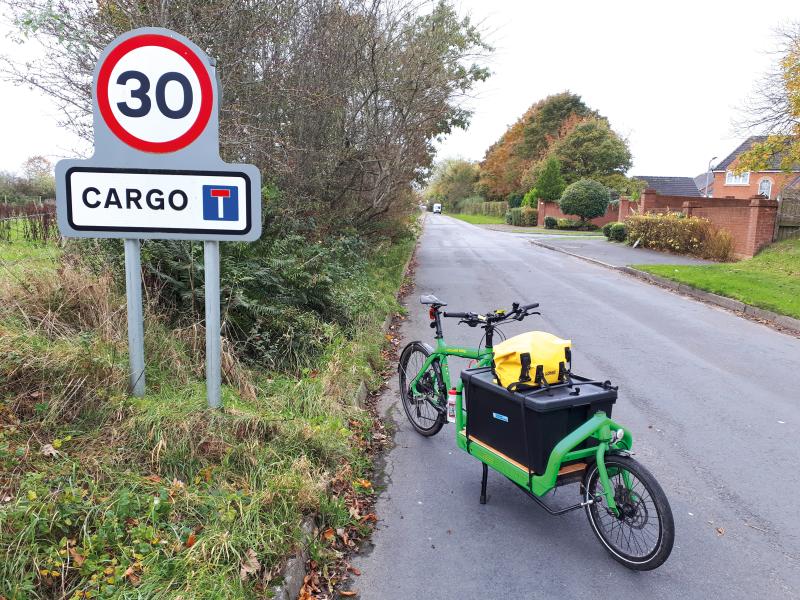 A cargo bike stands in a residential road loaded up with a crate and bag. in the foreground, a road sign reads '30' with the word 'Cargo' and the symbol for a dead-end underneath