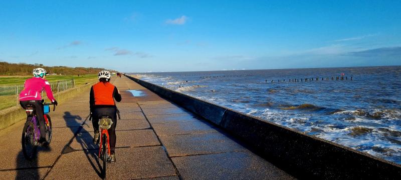 Two people cycle along a concrete sea wall in bright sunshine, with waves lapping the shore on their right