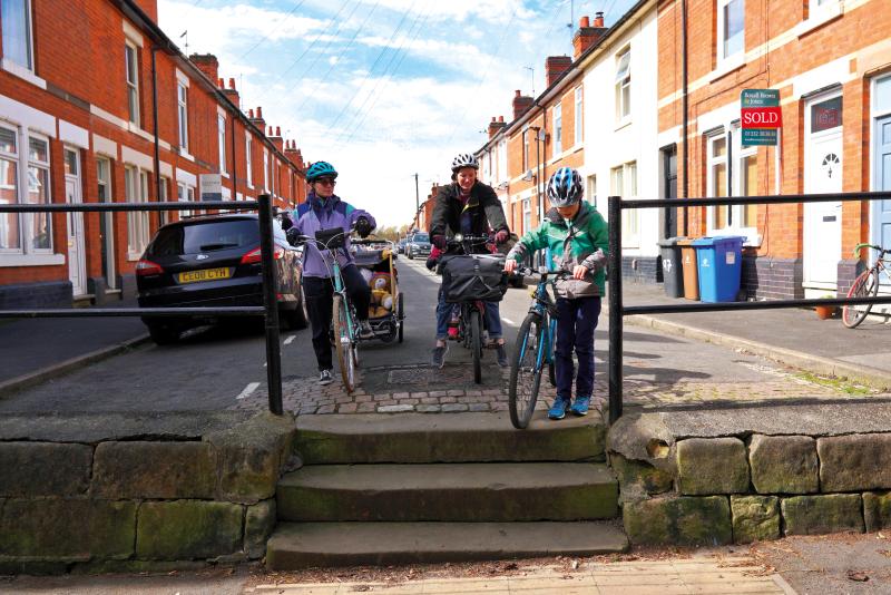 A child and two women, one of them towing a child trailer arrive at a set of steps at the end of a terraced street.