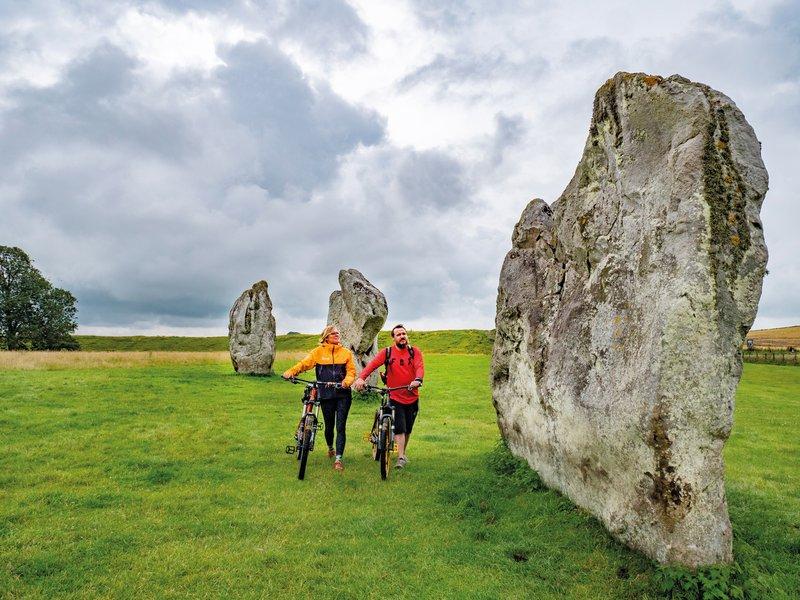 A woman and a man walk through a field dotted with stone monuments towering above them. They are both wheeling bicycles.