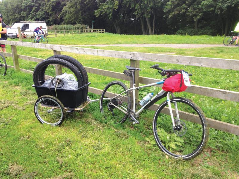 A bike is leaning against a fence. It has a trailer attached to the back which has replacement wheels for the bike