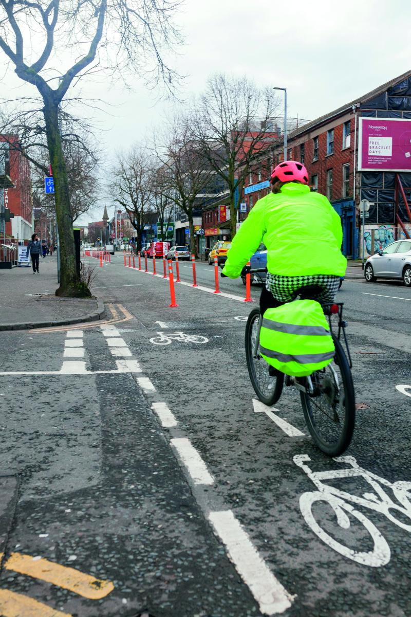 A woman cycles along a segregated cycle lane. She is wearing a helmet and high-vis and carries a pannier bag on her bicycle