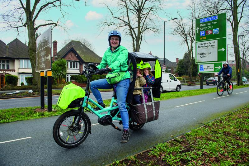 A woman pauses on a segregated cycle lane alongside a busy road. She is riding an e-cargo bike carrying two children under a covered shelter