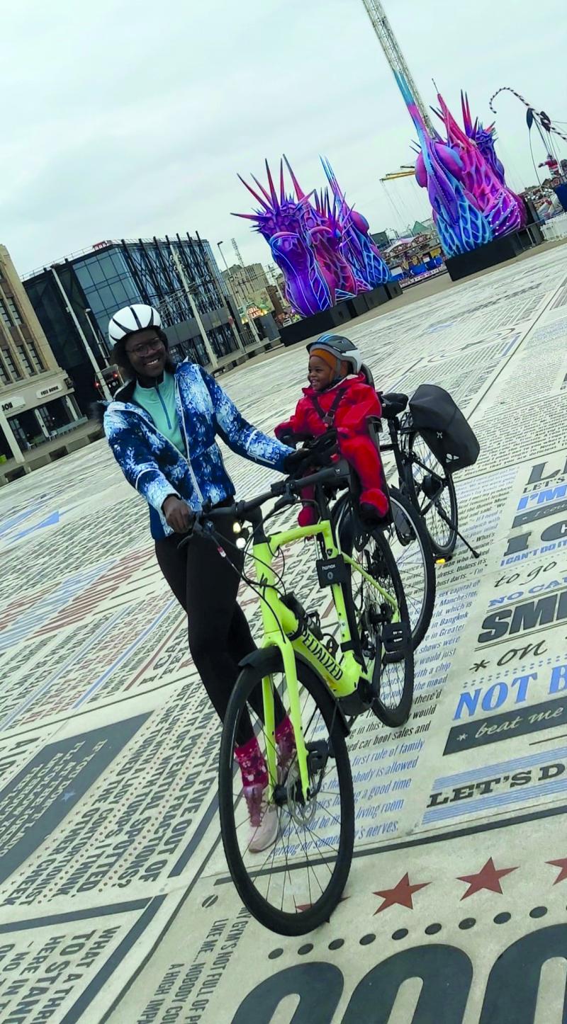 A woman stands holding her e-cycle. Her toddler sits in a child seat over the back wheel.