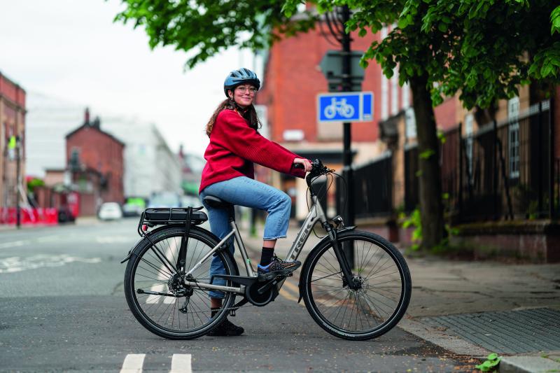 A woman poses side-on in a cycle lane along a quiet urban road. She is wearing casual clothes and smiling at the camera