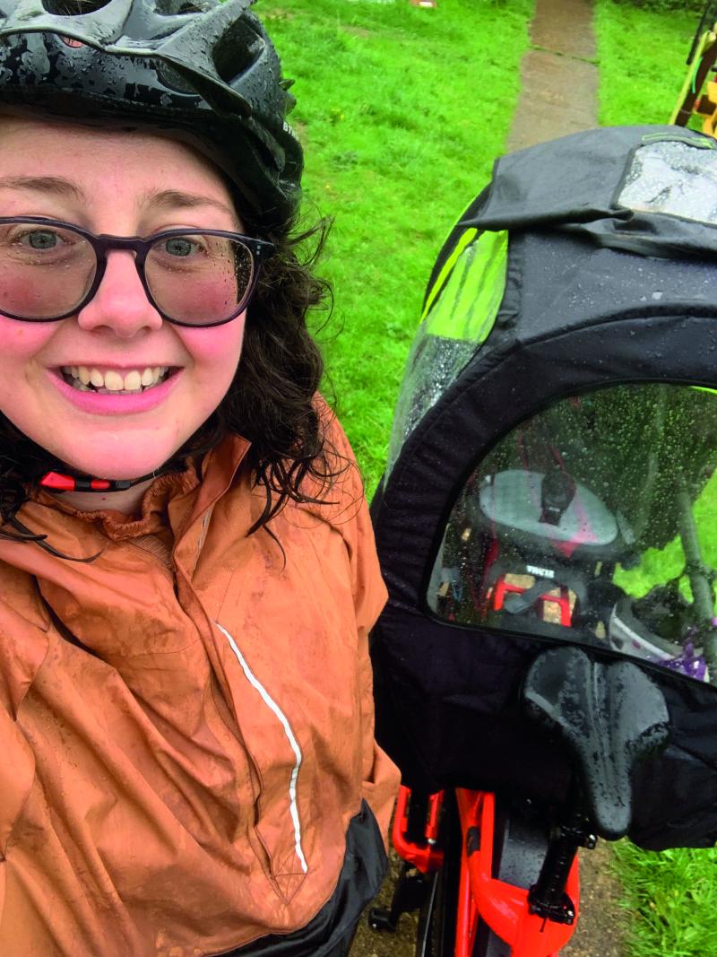 A woman smiles at the camera whilst taking a selfie. She is wearing a raincoat, helmet and glasses. Behind her is the e-cycle which has a rain shelter for the passenger compartment
