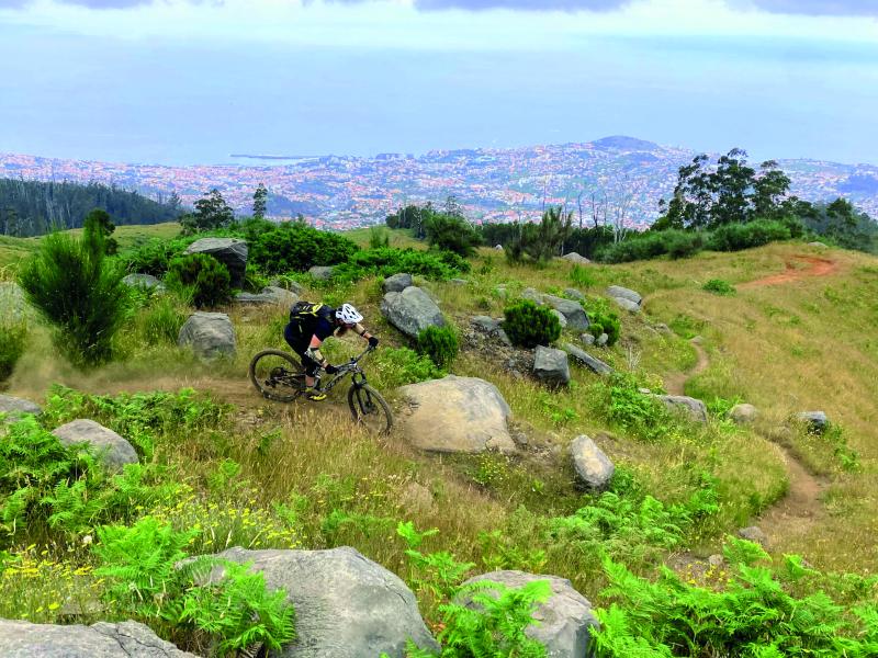 A mountain biker cycles along a dirt track through bracken. In the background a coastline is visible, crowded by red-roofed buildings