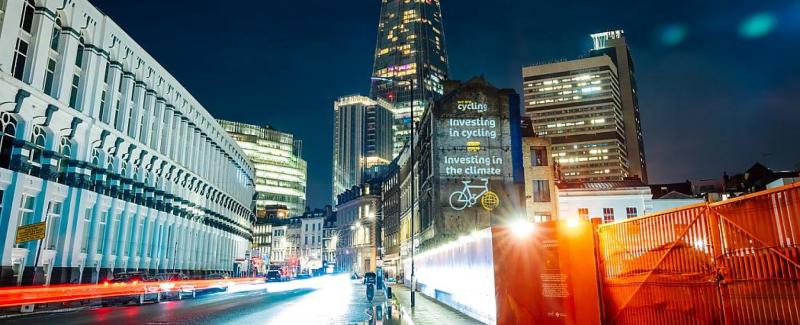 Night time photograph: the Cycling UK logo and a slogan saying &quot;This machine fights climate change&quot; are projected on the side of a building in London. The Shard skyscraper is in the background.