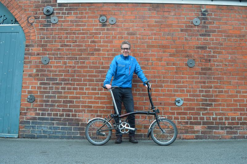 A white man stands against a red brick wall holding a black Brompton foldable bike