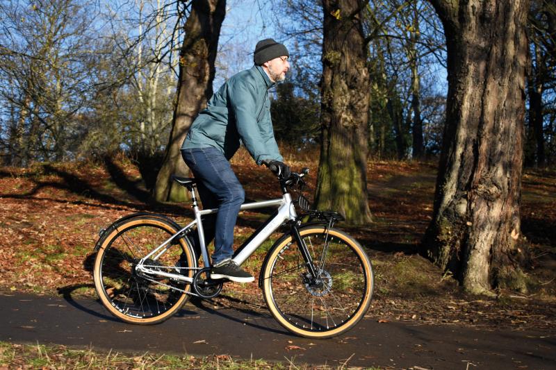 A man wearing a woollen beanie hat, gloves, jacket and jeans rides an e-bike through a wooded area on a tarmac path