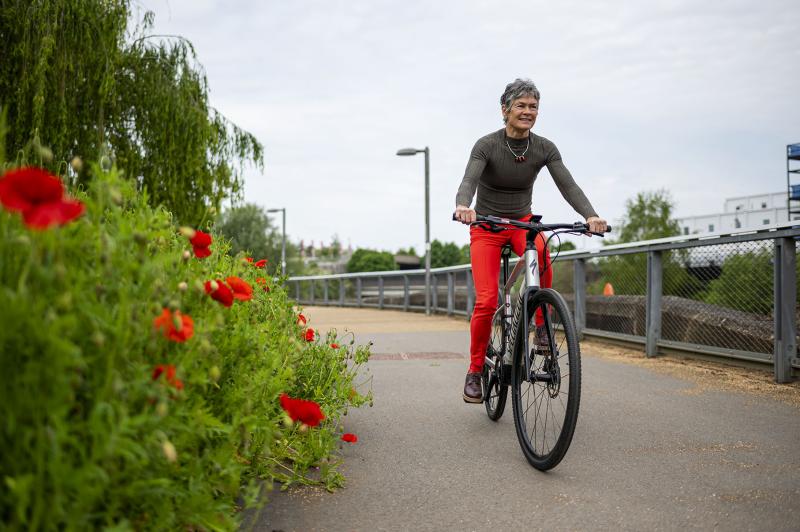 A woman wearing a grey long sleeved top and red trousers pedals beside a green bush with red flowers.