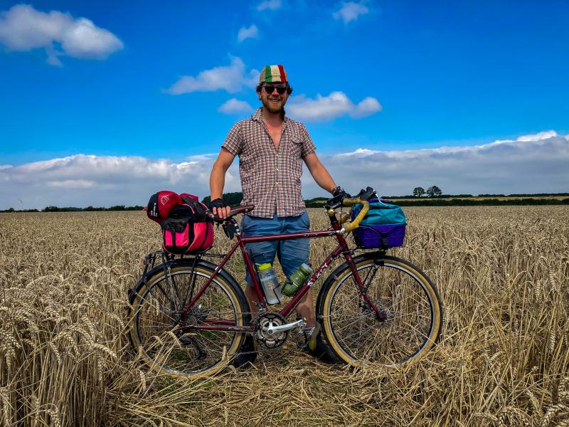 A man stands in a field holding his bicycle, It's a maroon coloured off-road bike with a bag in a basket on the front and a bag and a tent/sleeping back on the back pannier rack
