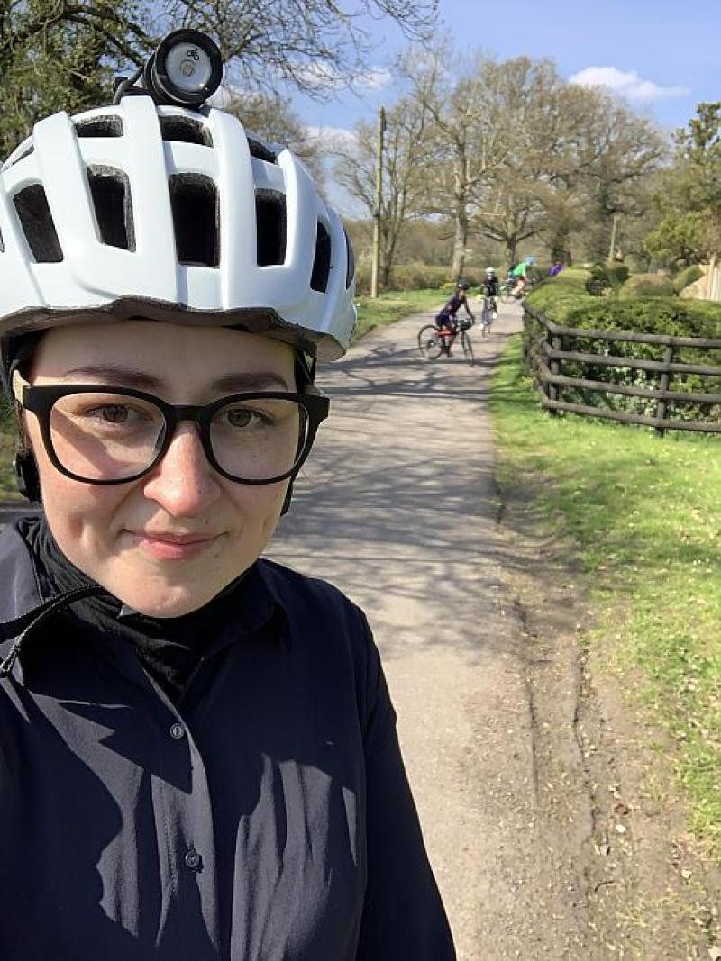 A woman wearing glasses and a helmet. She is posing for a selfie on a path ahead of other cyclists
