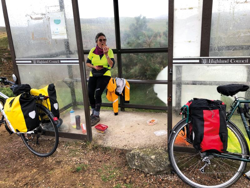 A woman sits in a bus shelter, her snacks and drink scattered on the floor, her bicycle perched against the outside of the shelter