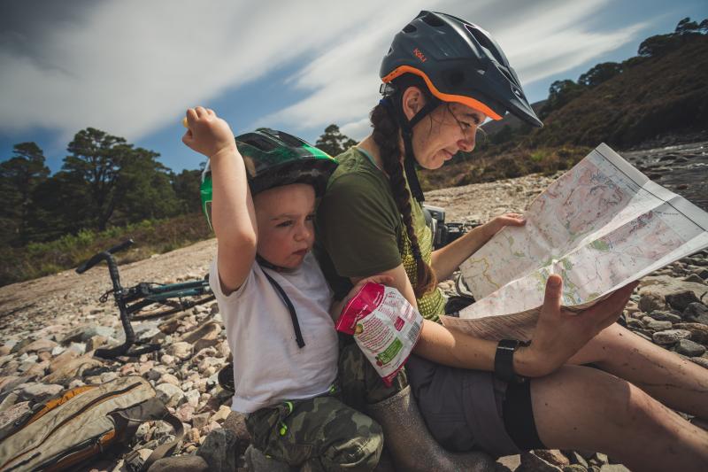 A little boy sits next to his mother who is reading a map. They are both wearing helmets and their bicyles lay behind them