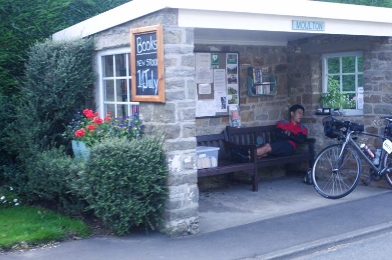 A tired rider sleeps on a bench under an outdoor shelter with information on the local area and shelves of books that are being sold