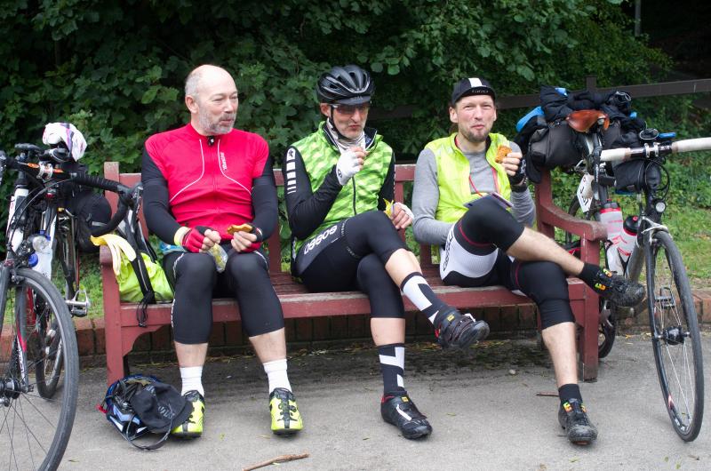 Three men are taking a well-earned rest stop, sitting on a bench and having a snack during the ride