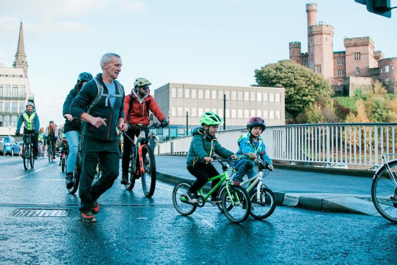 Children and adults taking part in a Kidical Mass ride in Inverness, with Inverness Castle in the background
