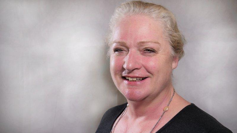 A woman with blonde/grey hair wearing a black top positioned in front of a white backdrop