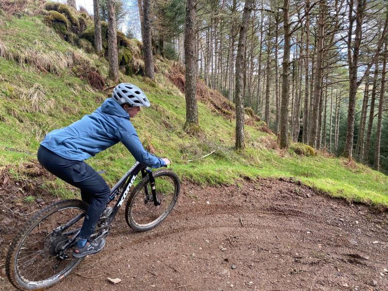 A women wearing a grey helmet, blue jacket and trousers rides a mountain bike downhill in Scotland