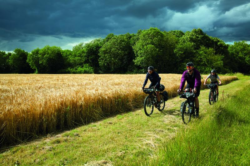 Three cyclists on off-road bikes, two female, one male cycle towards us along a grassy clearing in front of a stormy sky
