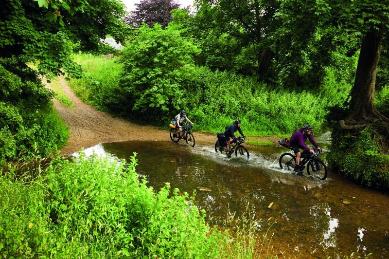 Three cyclists ride through what appears to be a sandy ford crossing a country lane