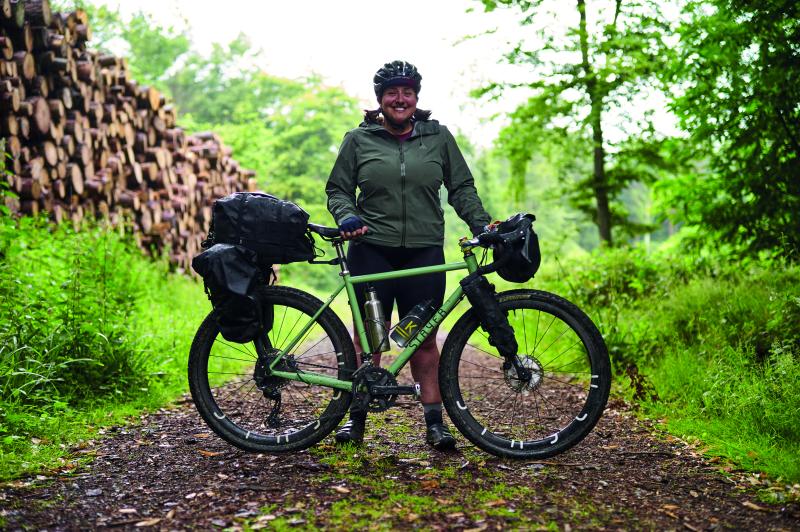 A woman stands on a wooded path behind her bicycle