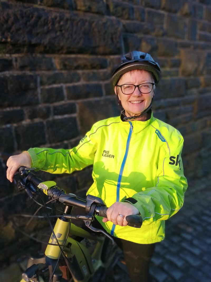 Kate Wylie with her bike, wearing a bright yellow hi vis jacket and a helmet, smiling at the camera