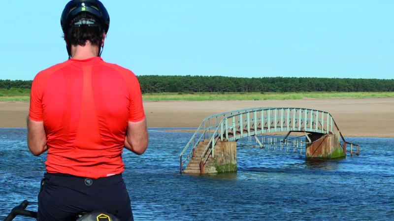 A man stands in the foreground, his back to the camera. He's wearing a bright orange jersey and a black helmet. In the background is a bridge crossing a river