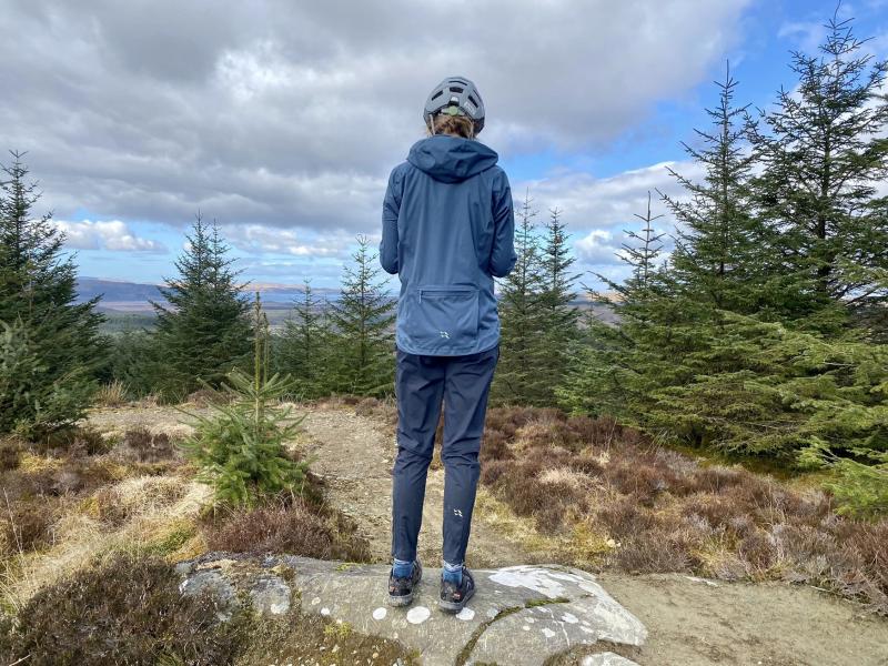 Jo stands at the top off a trail and looks at a map as she wears the Rab Cinder Kinetic waterproof pants and jacket