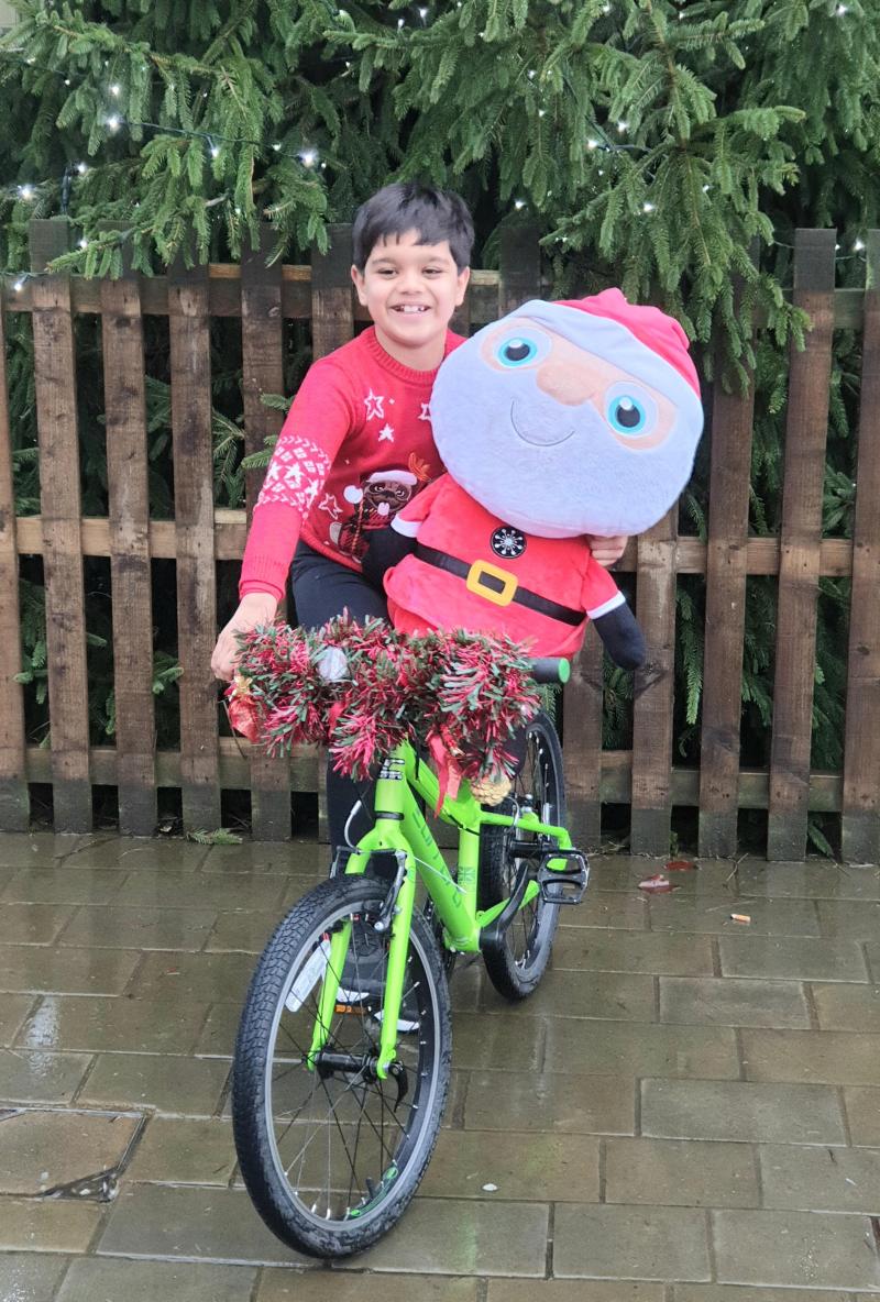 A young boy holds an inflatable Santa while sat on his bike against the backdrop of a wooden fence and a Christmas tree