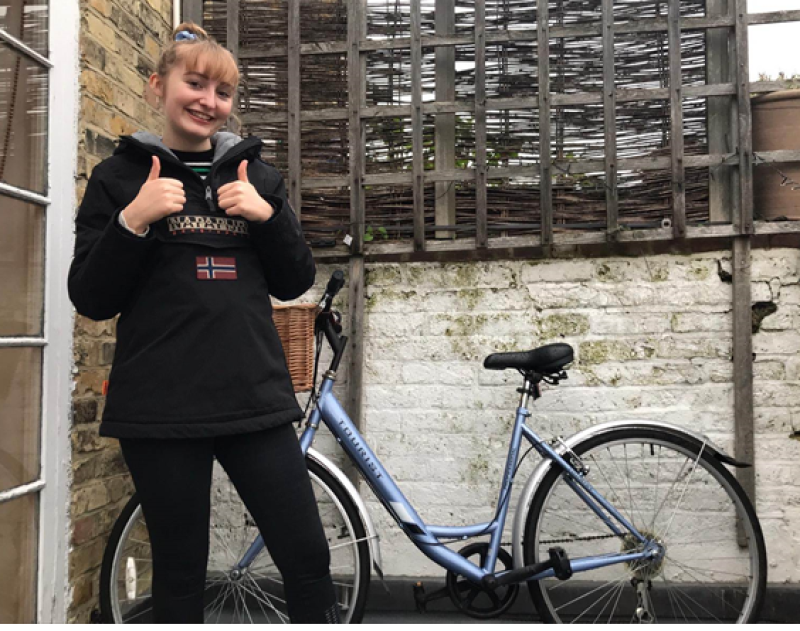 A young woman with blonde hair in a black hooded top poses with her bicycle in the background