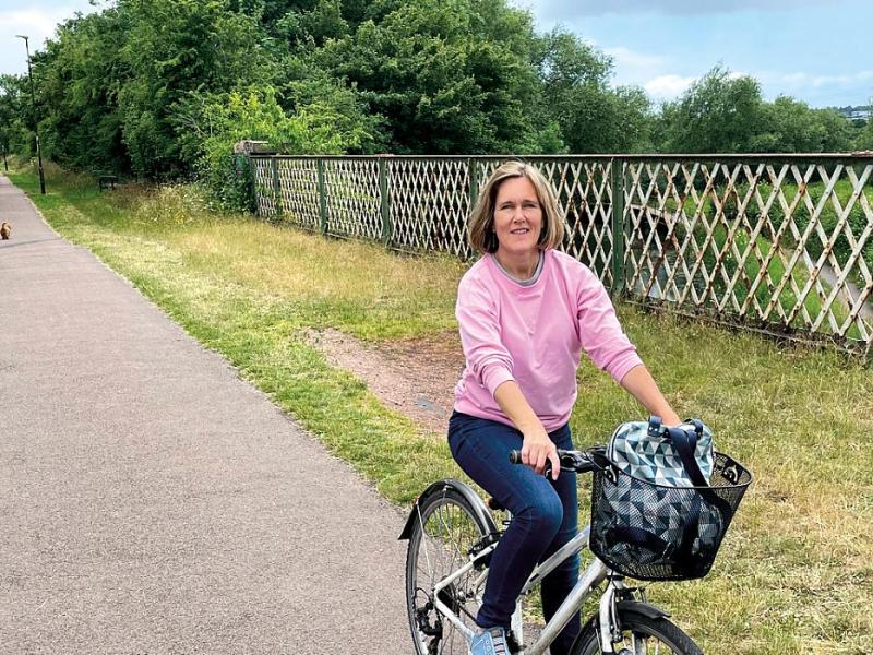 A blond woman cycles along bridge on a trail. She is looking at the camera and wears a pink jumper. She is not wearing a helmet