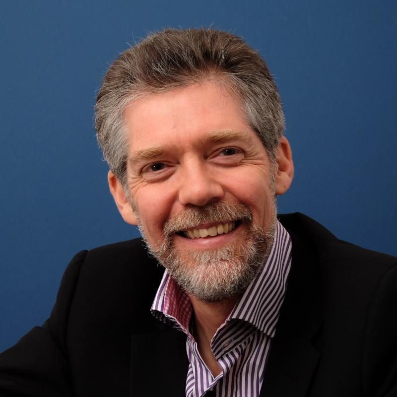 A middle-aged man with short hair and a beard wearing a dark suit and striped shirt