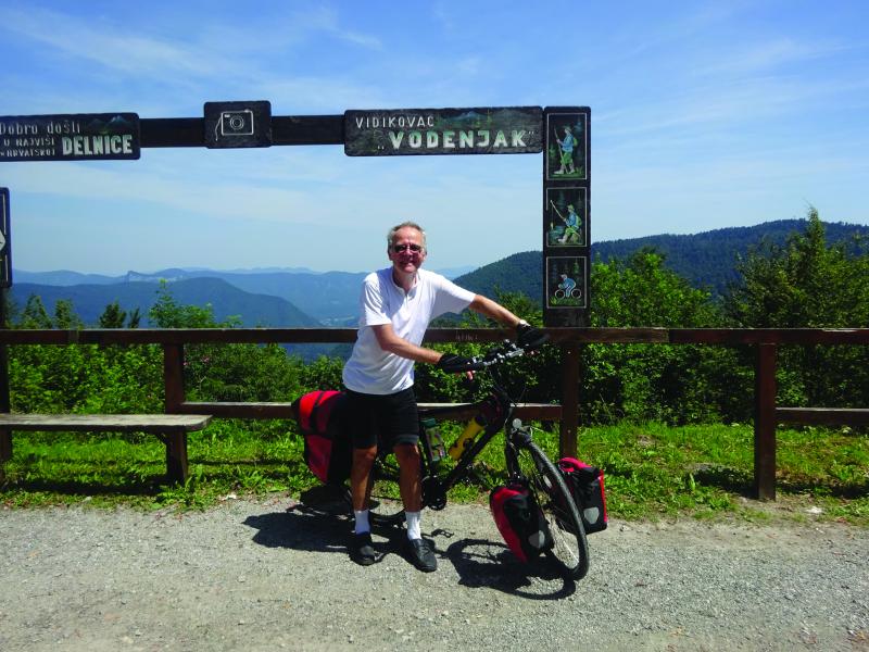 A man poses with his bicycle on a gravel track at the top of a mountain