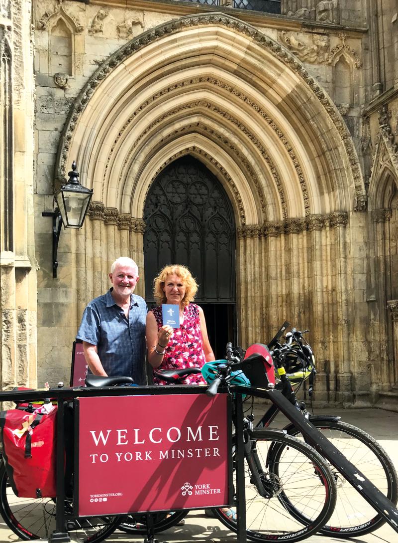 A man and woman stand with their bicycles in front of the large doors to a cathedral, a sign in front of them reads 'Welcome to York Minister'