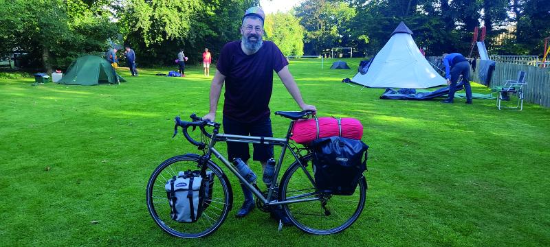 A bearded man stands on a campsite holding his bicycle which is loaded down with panniers